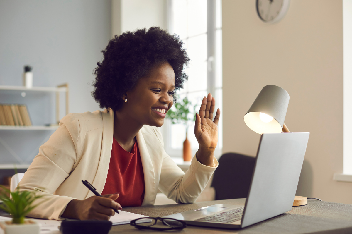 A woman sitting at a desk waving to her laptop and taking notes